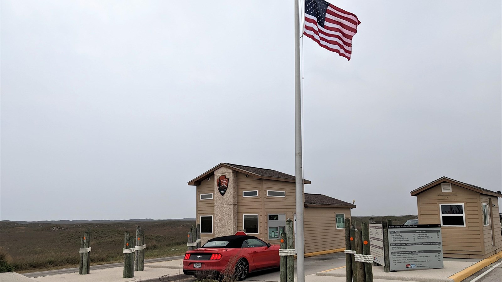 A red car drives up to a small brown building. An American flag flies overhead. 