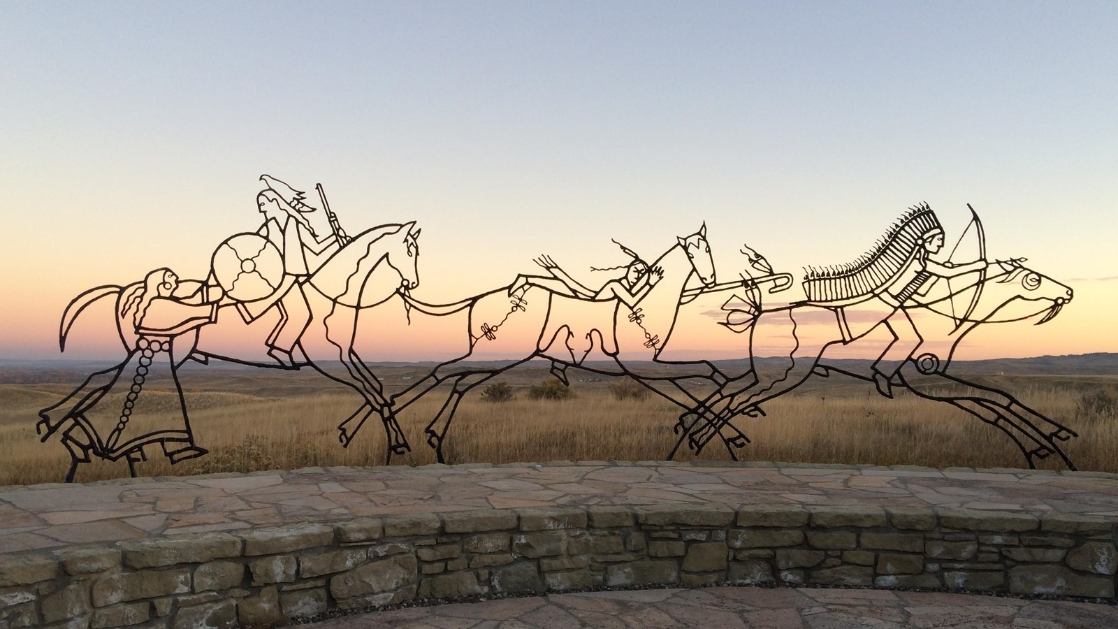 A large black metal sculpture of 3 Warriors and a woman on Horseback with sunset in background