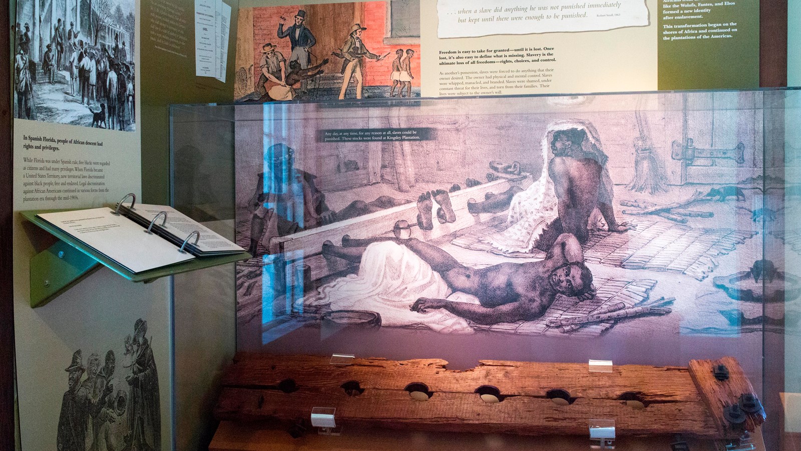 An exhibit case with wooden legs stocks and image behind shows illustration of enslaved man laying o
