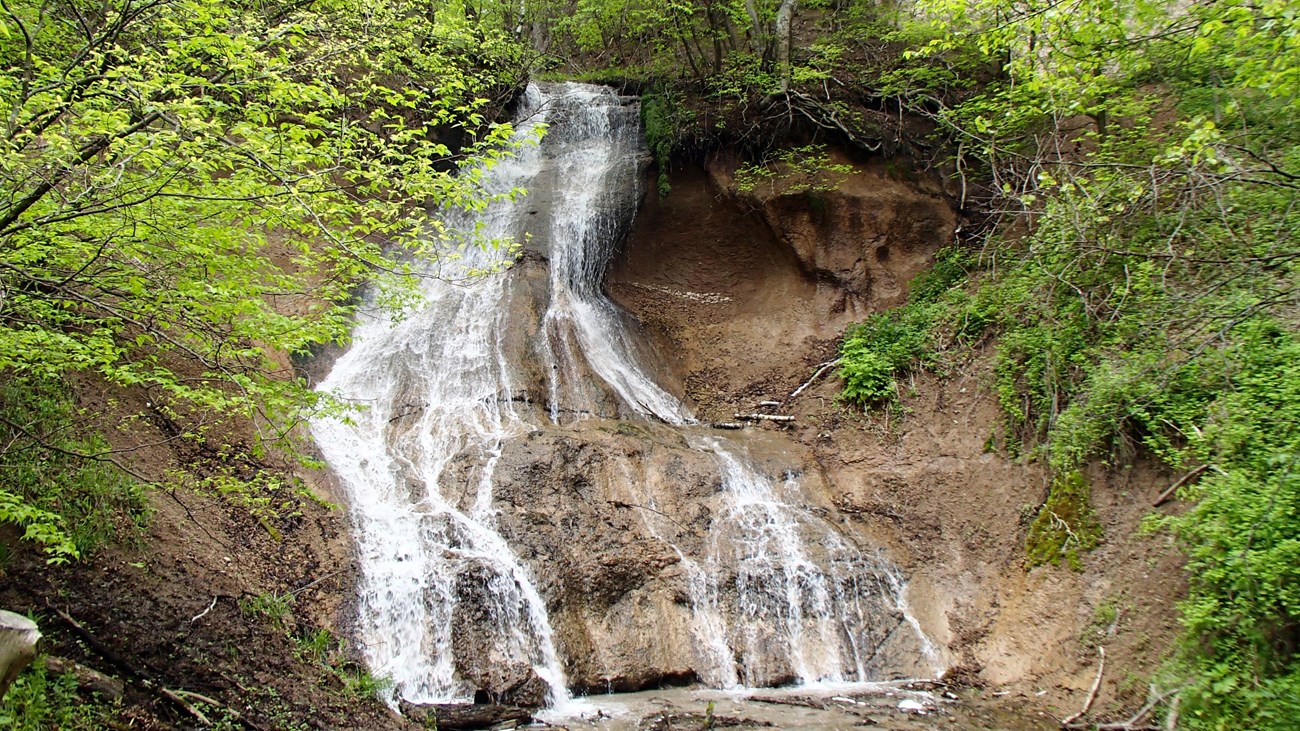 A waterfall curves down the side of a dark canyon wall before forming a wide shallow pool below.