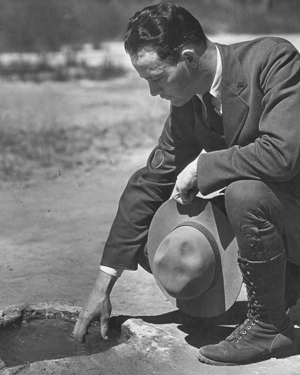 A black and white photo of a ranger crouching, dipping a hand into a small pool of water.