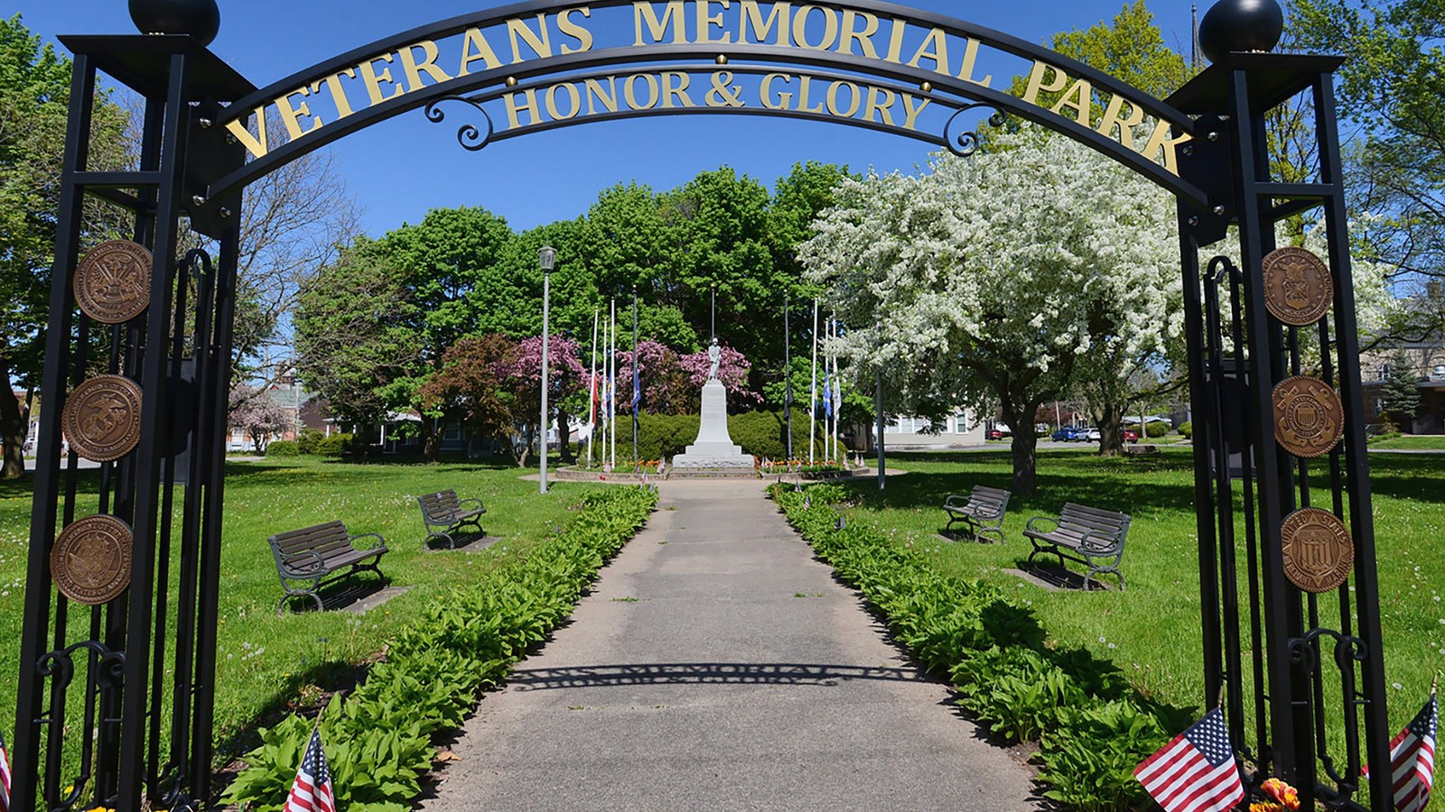 An iron arch at the edge of a sidewalk and grass. A statue stands in the distance beyond it. 