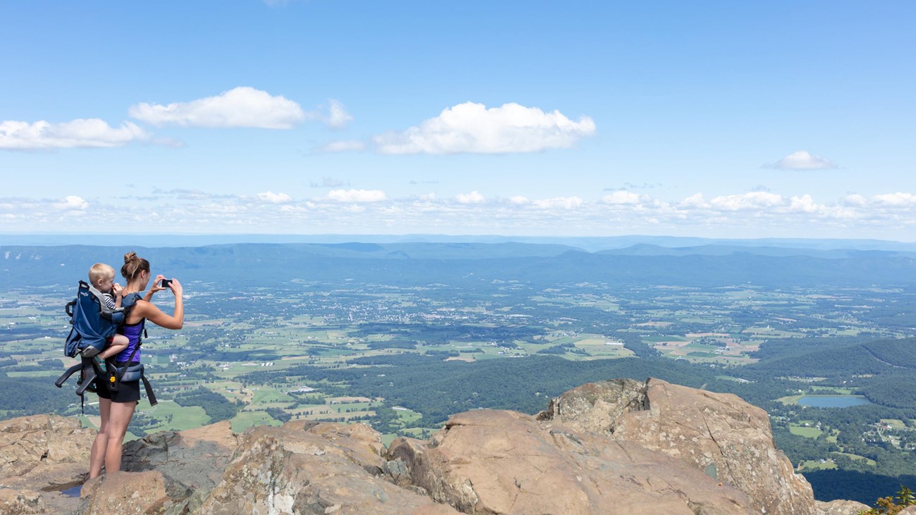 A woman with a baby on her back takes a picture with her phone on top of a mountain summit.