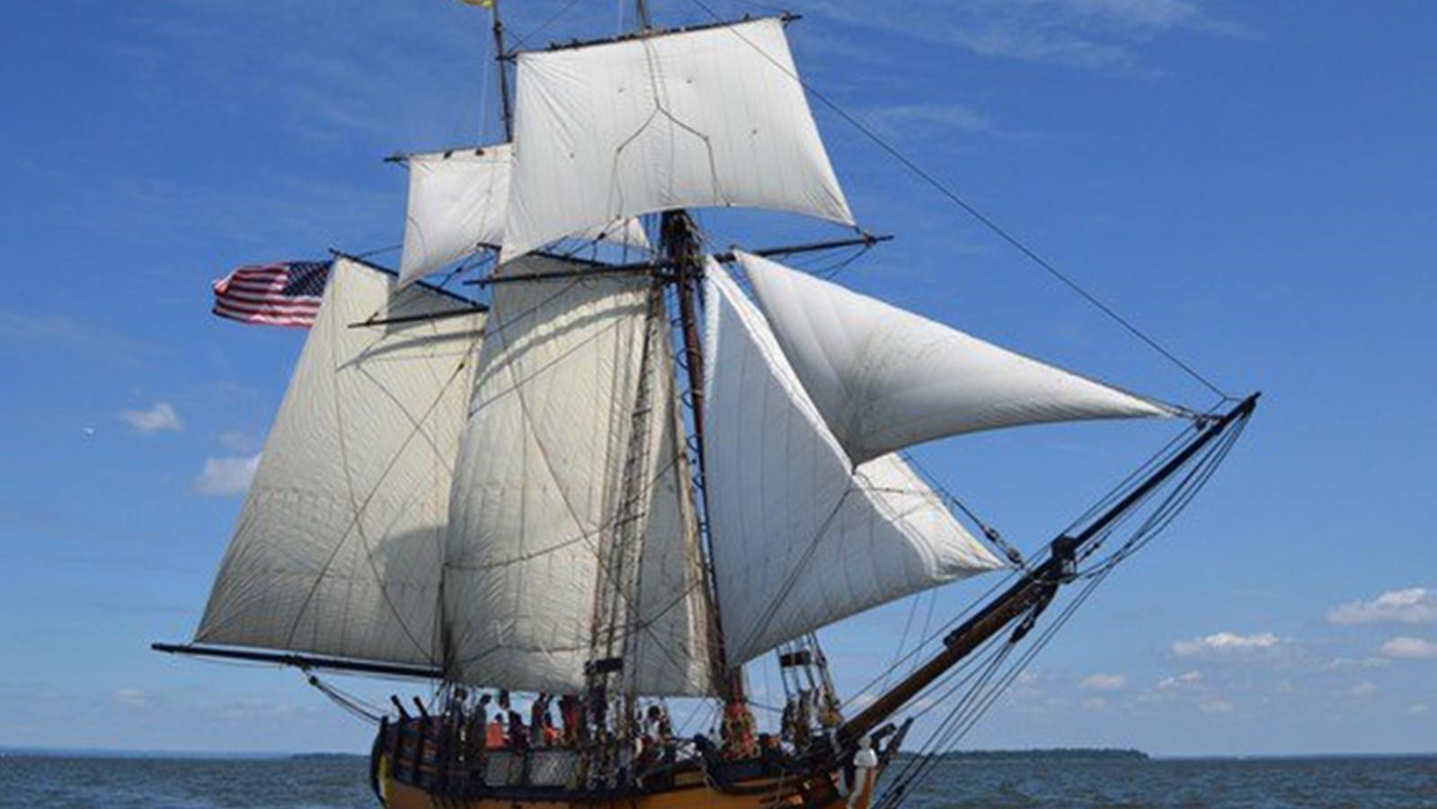 A classic sailing ship with two masts and white sails sailing on calm waters under a blue sky.