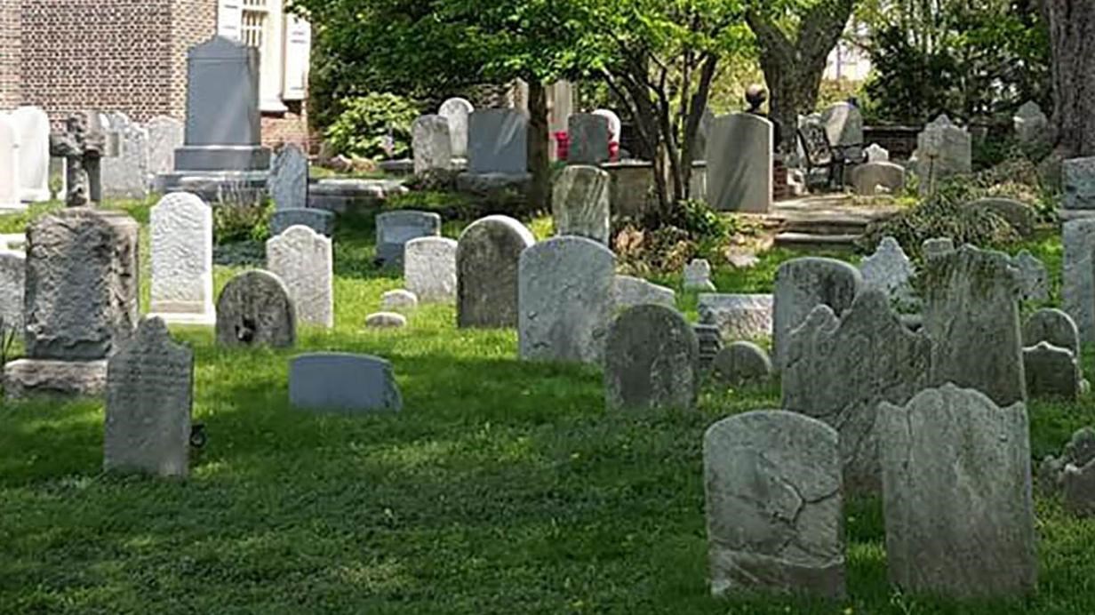 Headstones are scattered across a pastoral churchyard.  The brick church sits behind them.