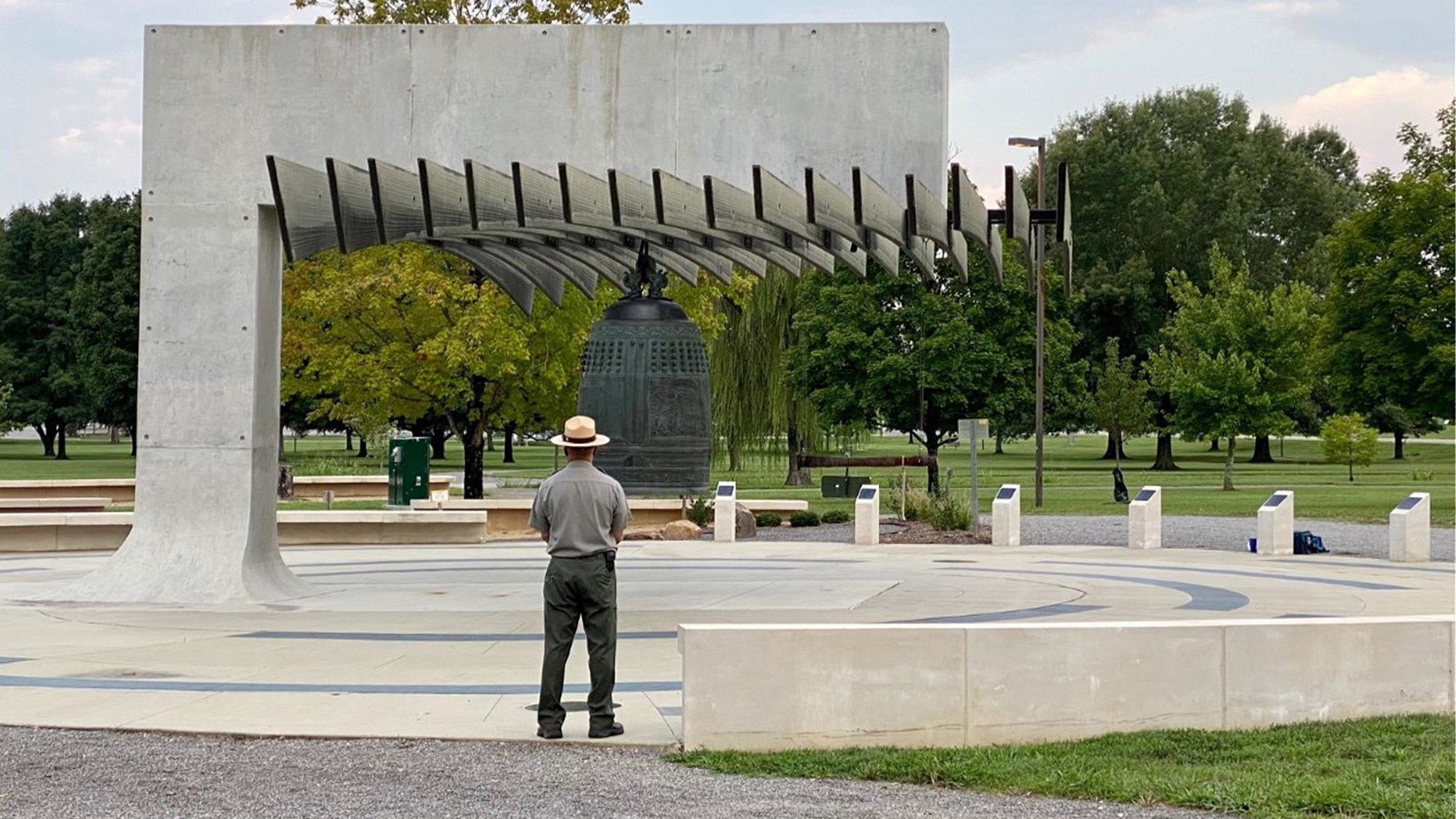 A person stands in front of a large bronze bell hanging from a  angled piece of concrete