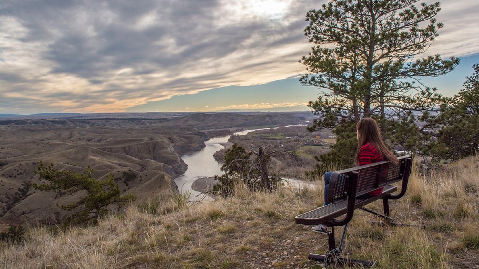 A woman sits on a bench overlooking bluffs and a river below
