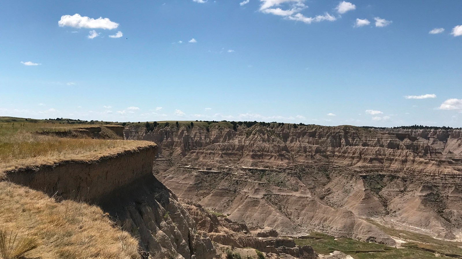 Sod layer dramatically falls over into badlands rock with a badlands table in horizon under blue sky