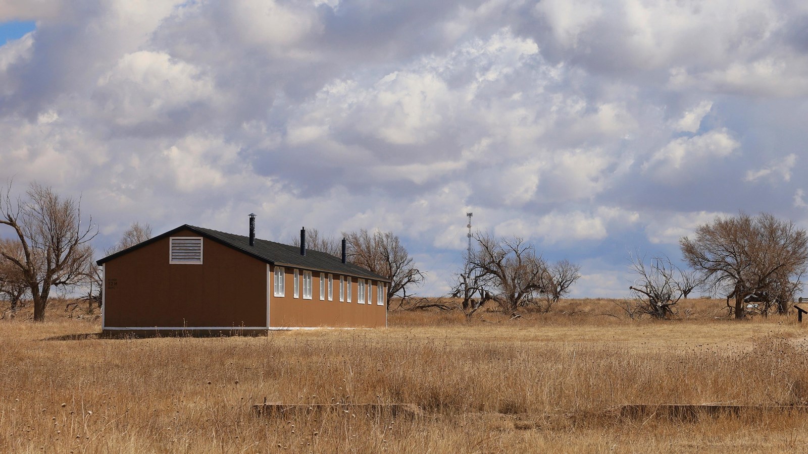 A brown grassy field under a cloudy sky, with a single story building and two water towers.