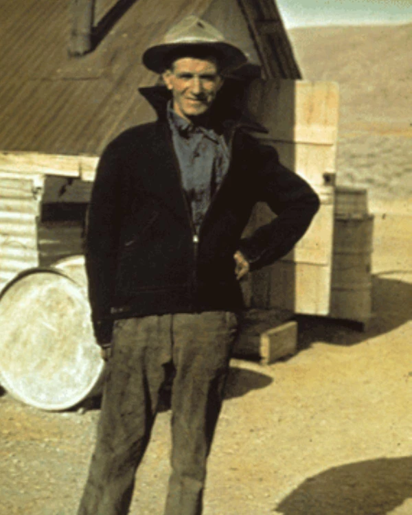 Man stands in front of house at a mining camp.