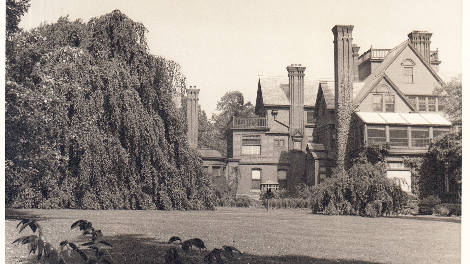 Black and White photo of a lawn and the back of a large home, with sky and trees surrounding.