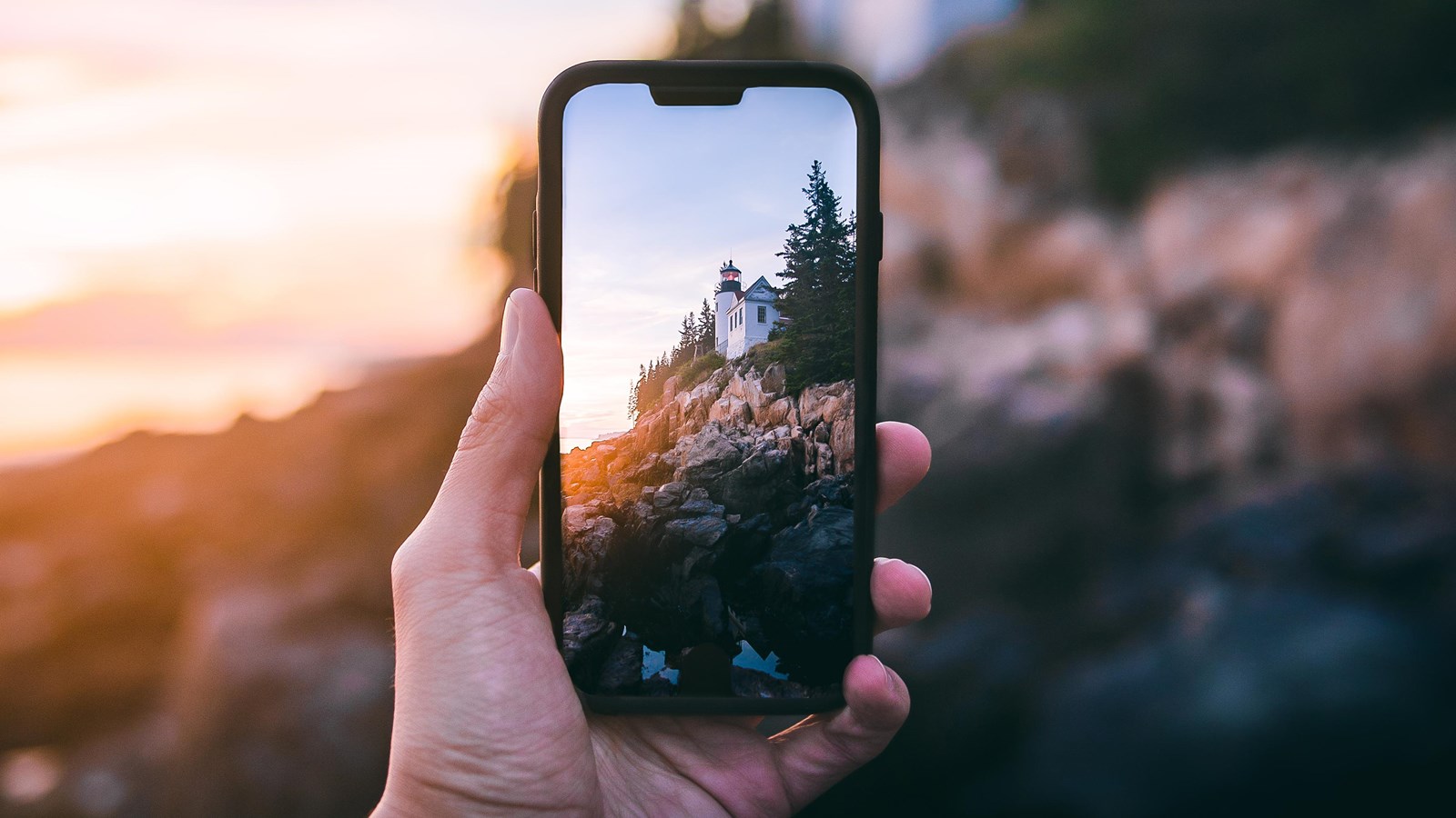 A hand holds a phone poised to snap a photo of a lighthouse bathed in sunset on a rocky coastline.