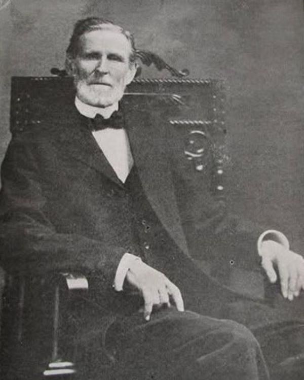 A man in a dark suit and bowtie sits in a chair.