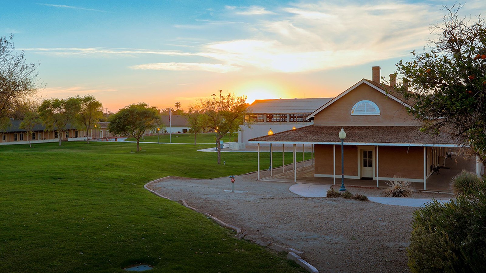 A view of an adobe structure with wrap around porch, manicured lawn and sunrise