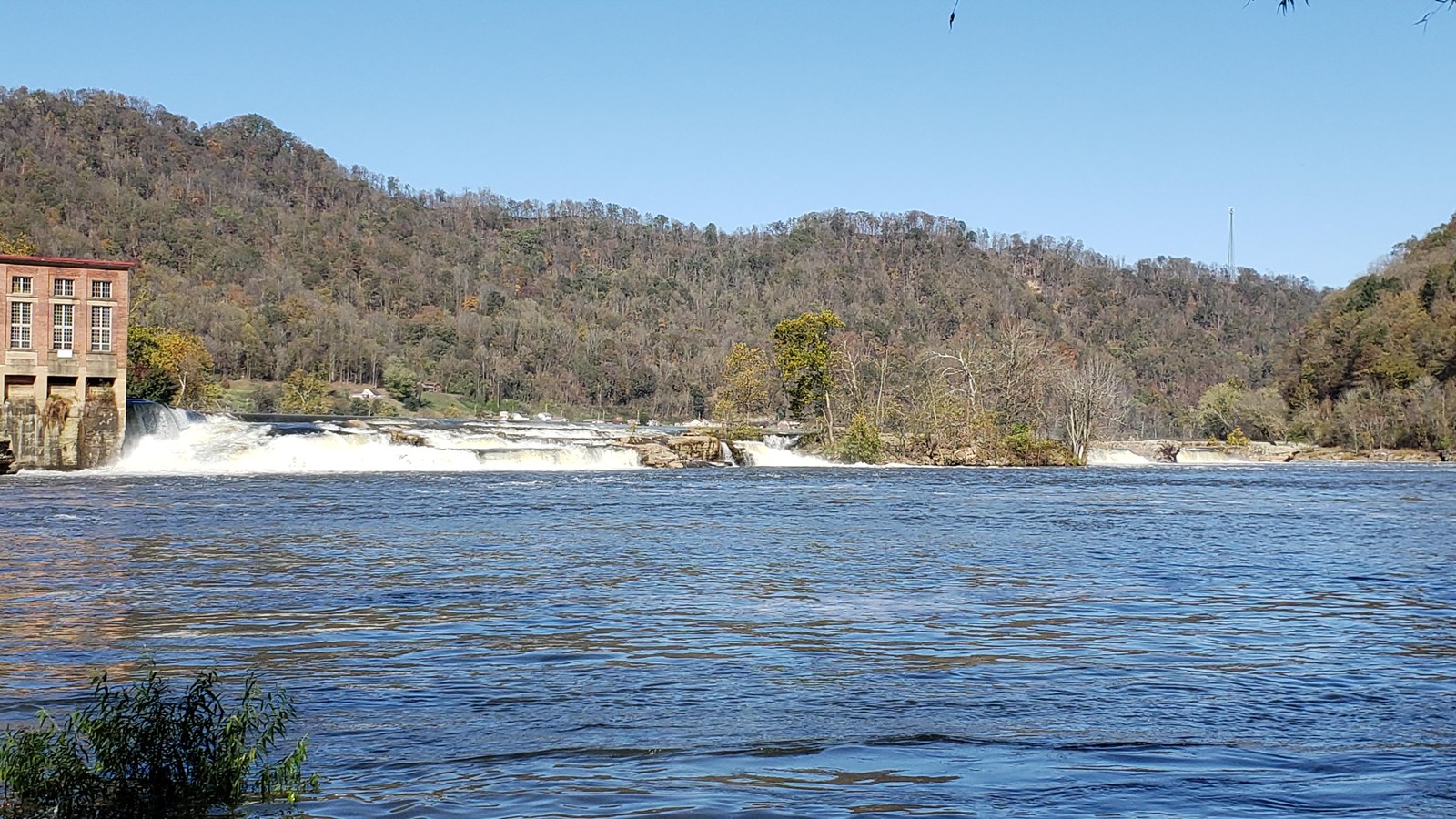 A large river flowing over sandstone rocks with a brick building on the shore surrounded by trees.