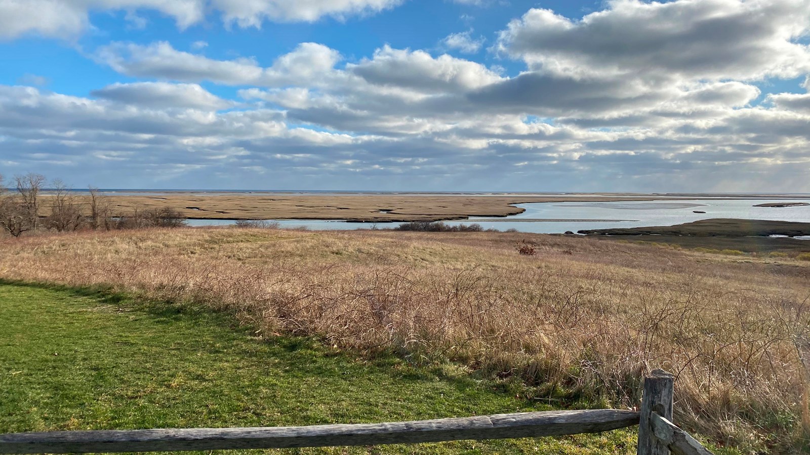 Grasslands from Fort Hill slope down to Nauset Marsh with a blue clouded sky.