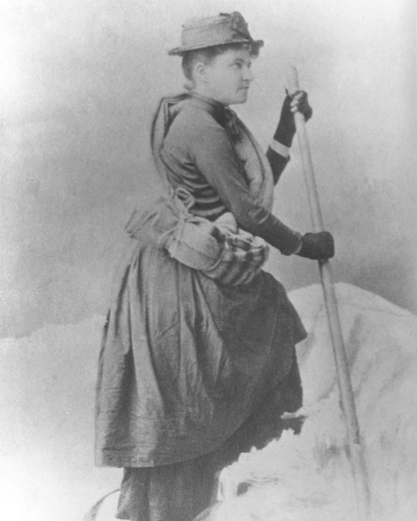 Black and white photo of a women posing with a blankets tied around her waist holding walking stick