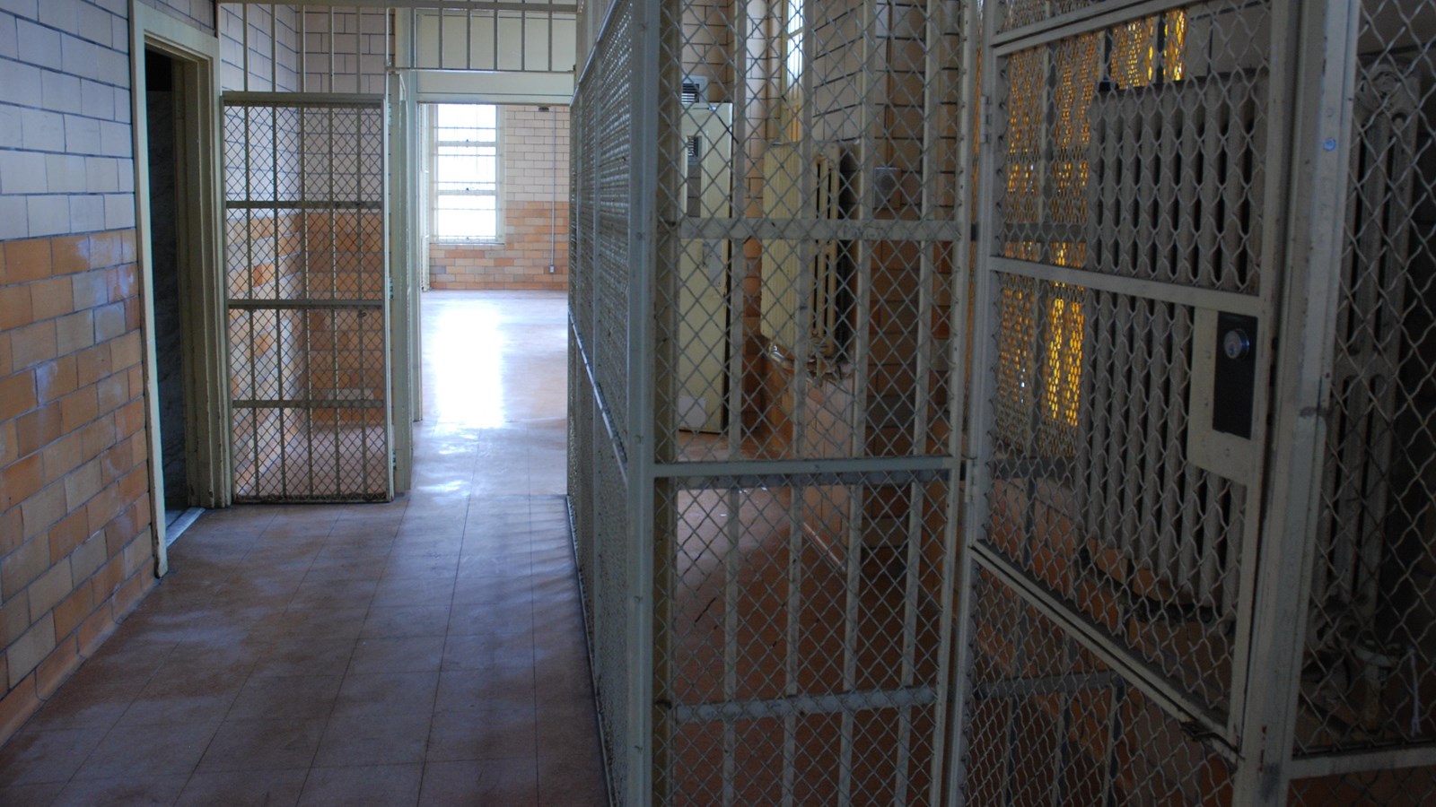 It features a corridor flanked by metal mesh screens and brick walls, illuminated by sunlight.