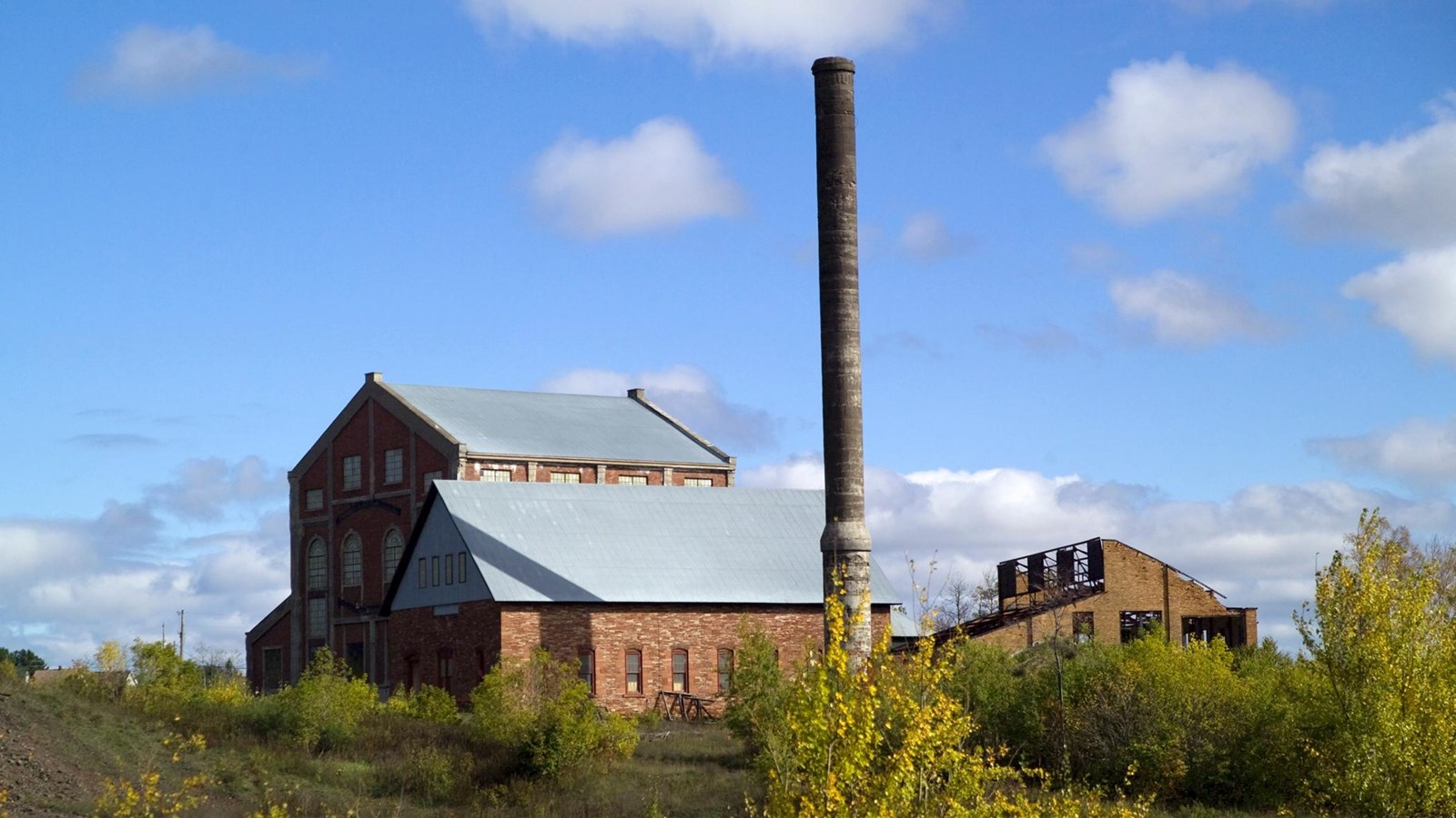 Three brick buildings, one in ruins, surround a tall smokestack.