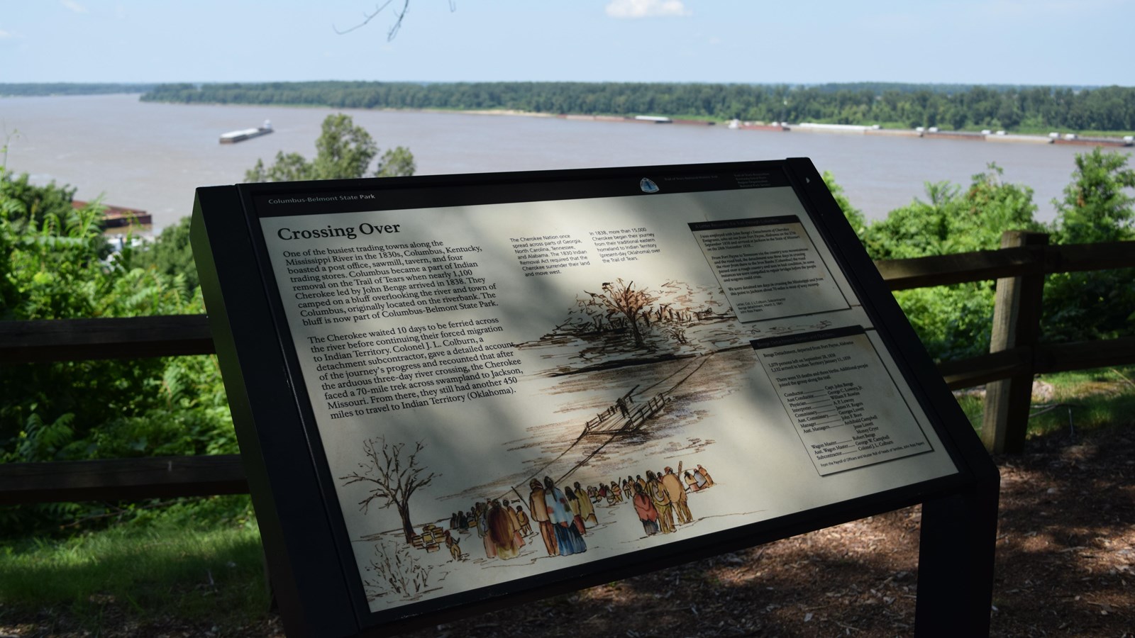 A sign sits next to a path overlooking a broad, flat river.