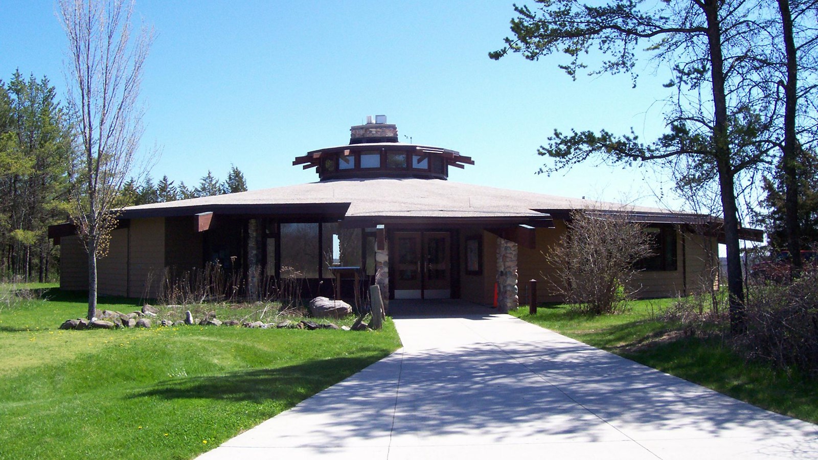 Round brown building surrounded by forest and green lawn with a paved path leading to its doors.
