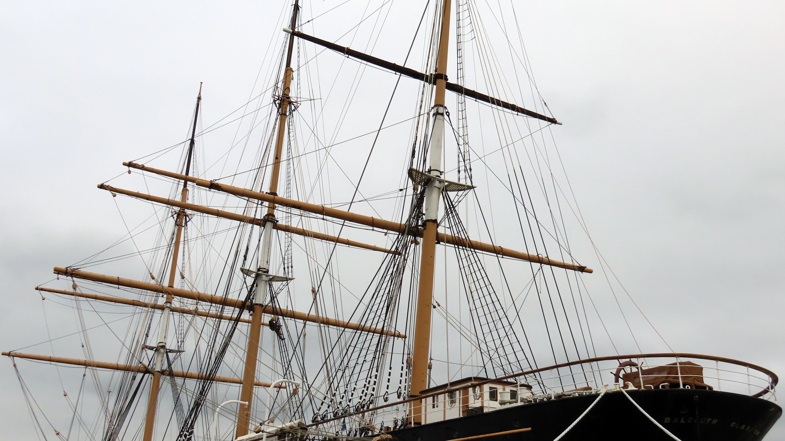 Three wooden masts with yards and rigging rising up from the deck of Balclutha, as seen from shore.