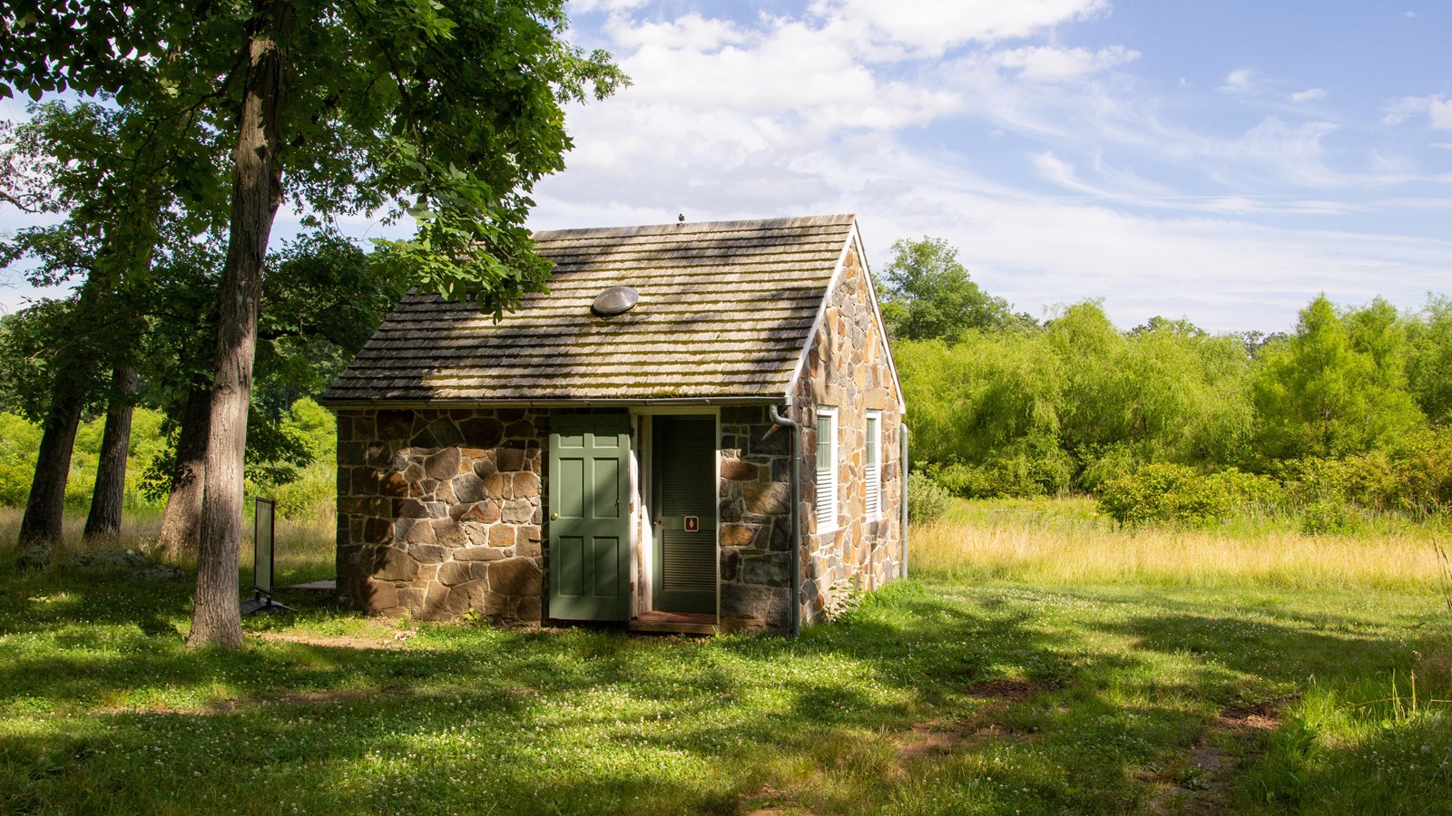 A single room stone building with a single door and a-frame roof surrounded by vegetation.