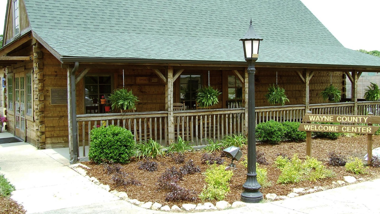 A log cabin looking building with a green roof stands with a landscaped front and porch.