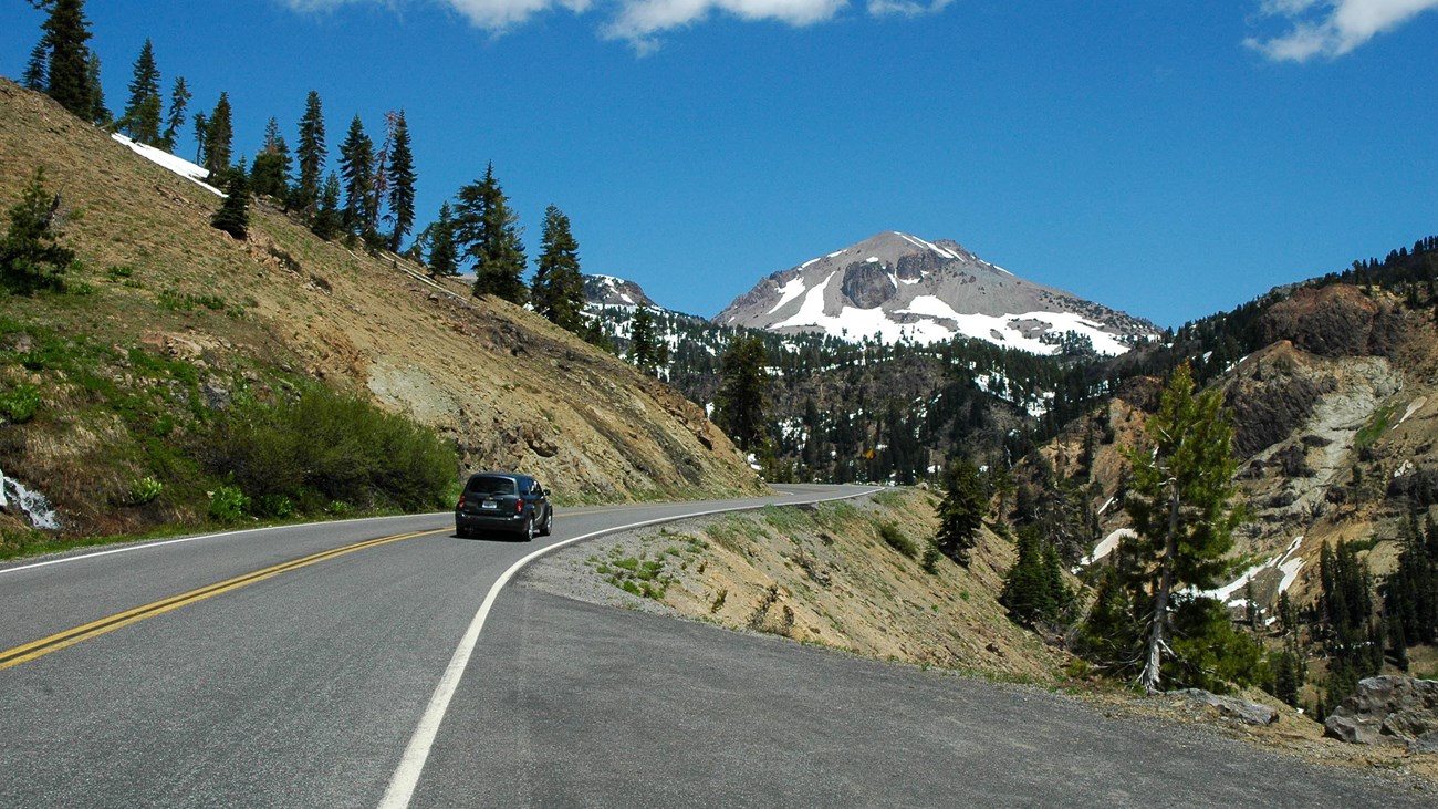 A black vehicle drives on a mountain road toward a large volcanic peak with large patches of snow.