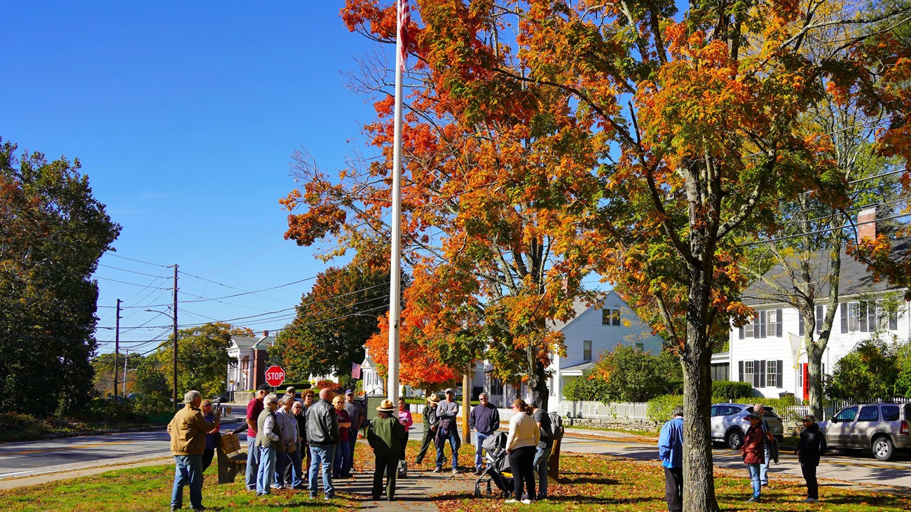 A ranger in NPS uniform stands under a flagpole surrounded by fall foliage speaking with visitors