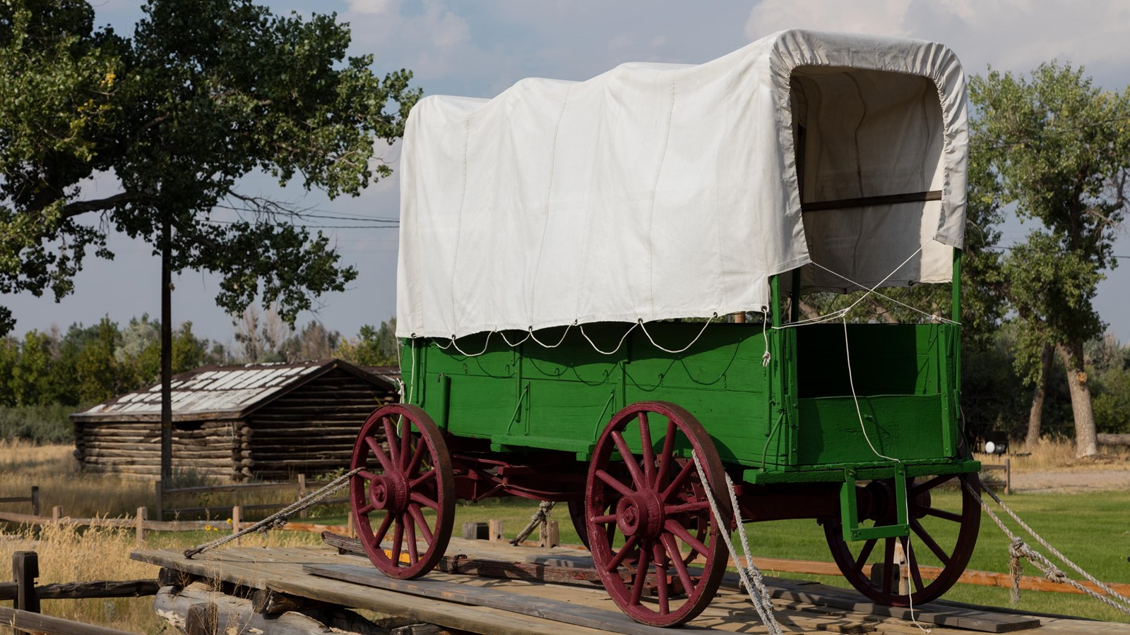 A large green covered wagon in front of a log cabin