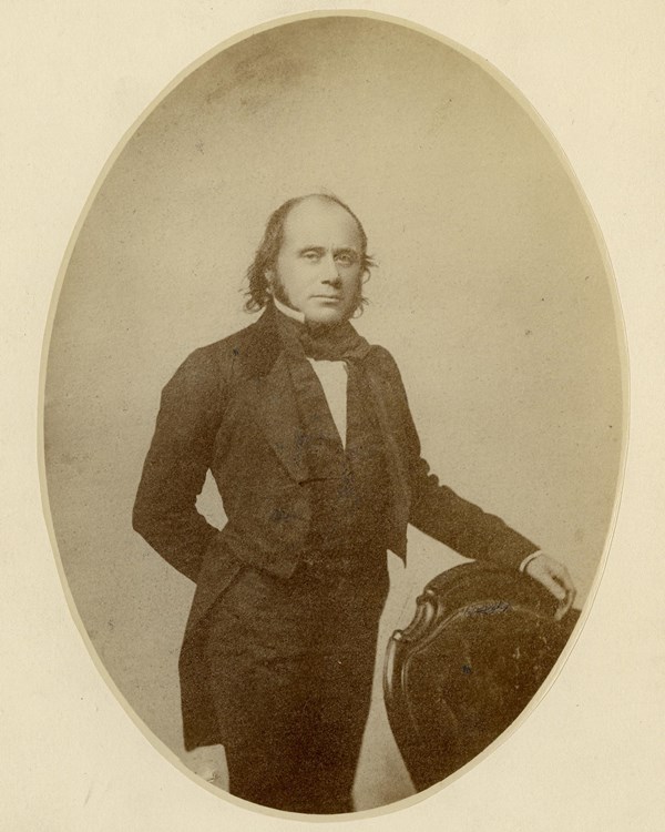 Man poses with his hand resting on the top of a chair. He wears a dress coat with a high collar.
