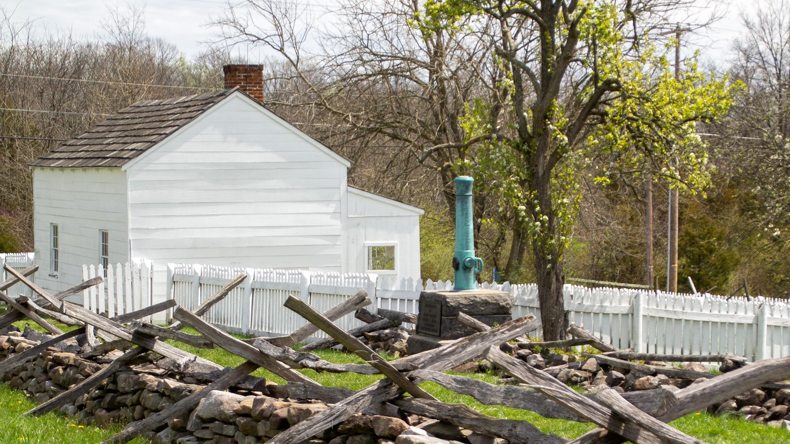 Small one story white barn near a rock & wooden pig tight fence and a teal colored canon monument.