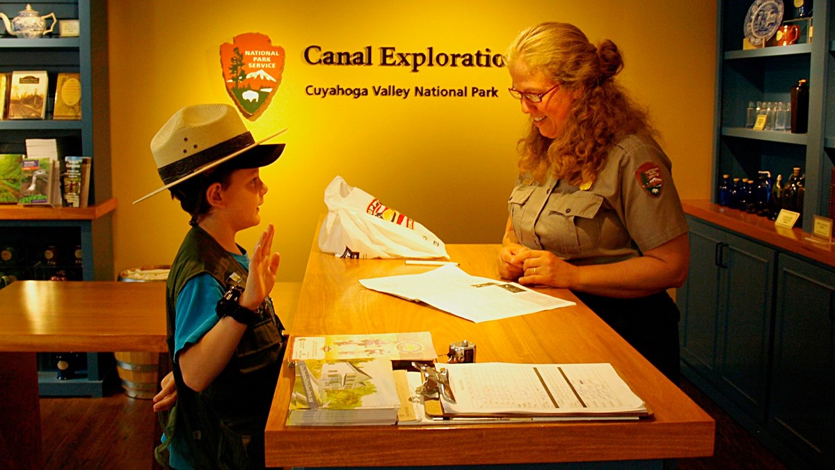A child wearing a ranger hat holds up their right hand, across a desk from a uniformed ranger.