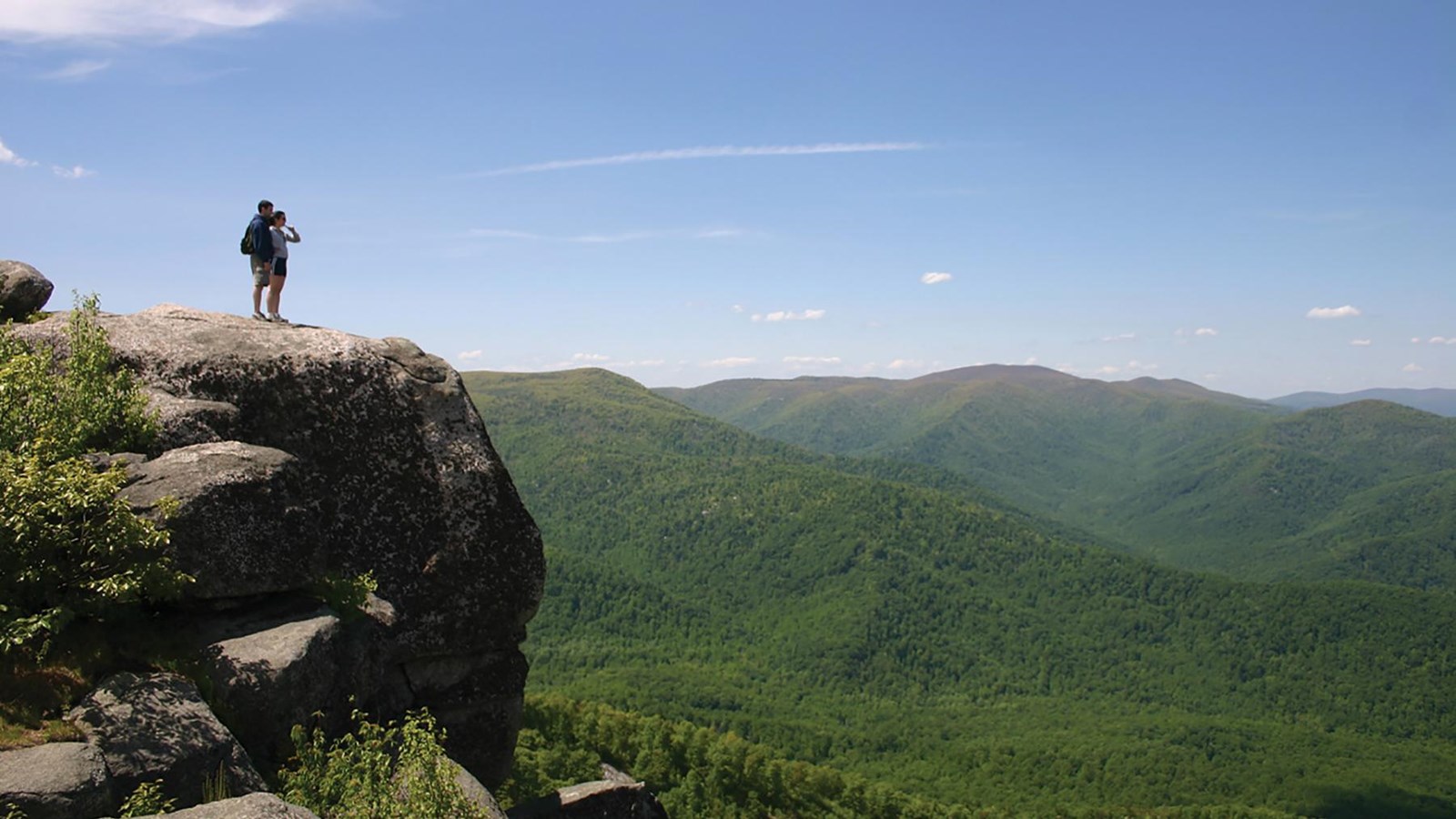 Two hikers standing on a huge boulder looking over a view of mountain ranges and a green valley