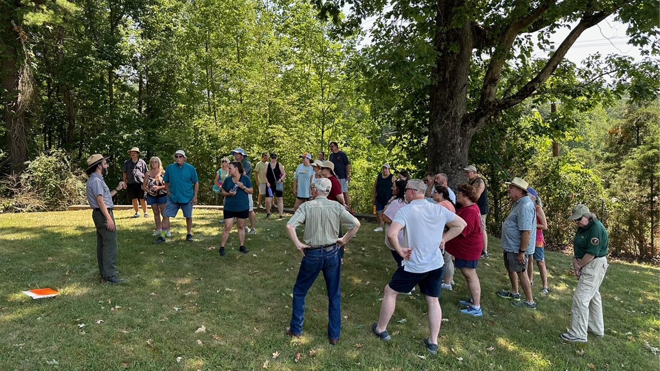 A ranger and several visitors gather on a grassy knoll surrounded by woods.