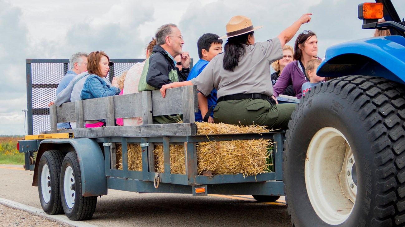 A woman in a flat hat and grey shirt points to an area over people\'s heads on a hay ride