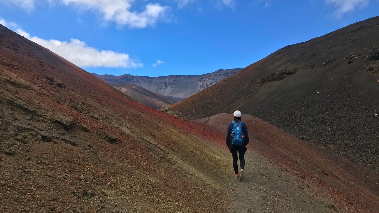 A hiker walks a trail between two red and yellow cinder cones. 