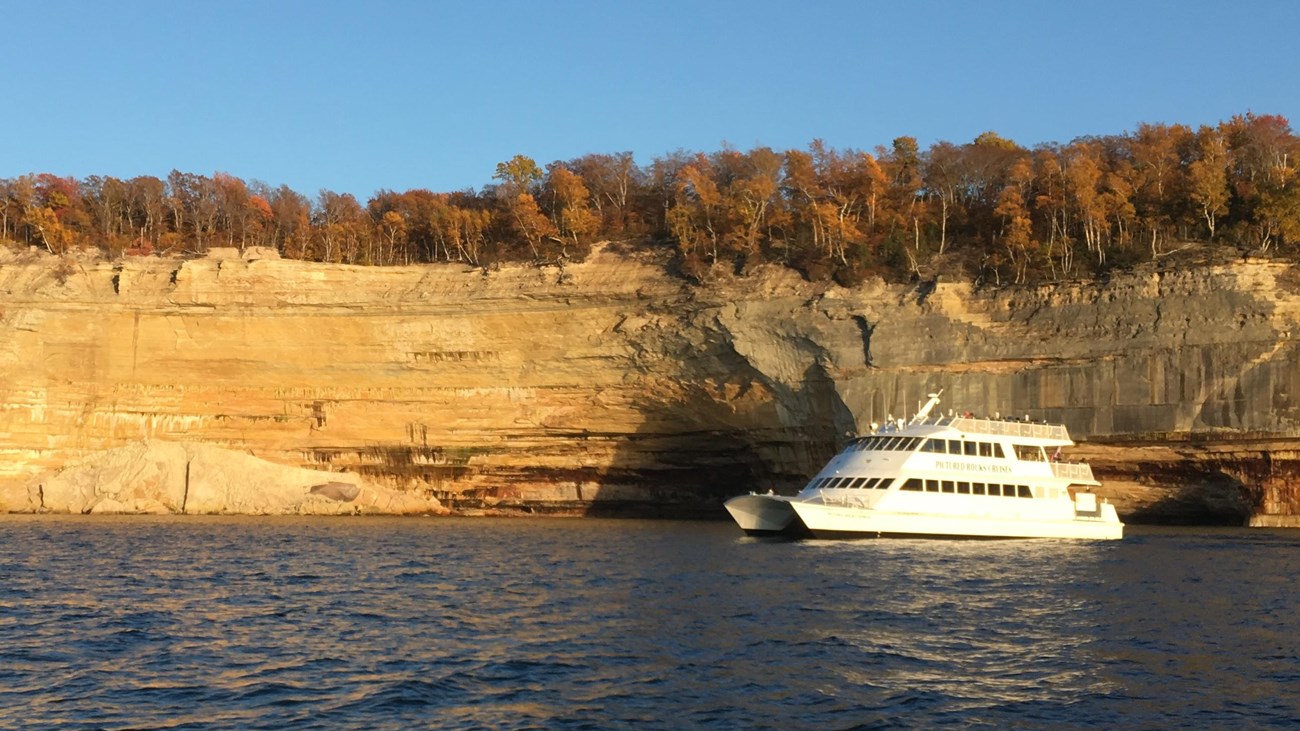 White cruise boat along cliffs with autumn trees on top.