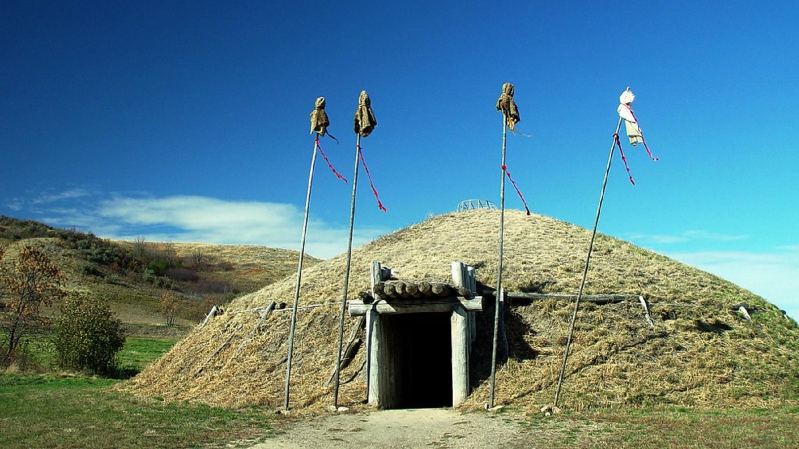 A domed earth structure covered in grass with a door going inside and poles around the door