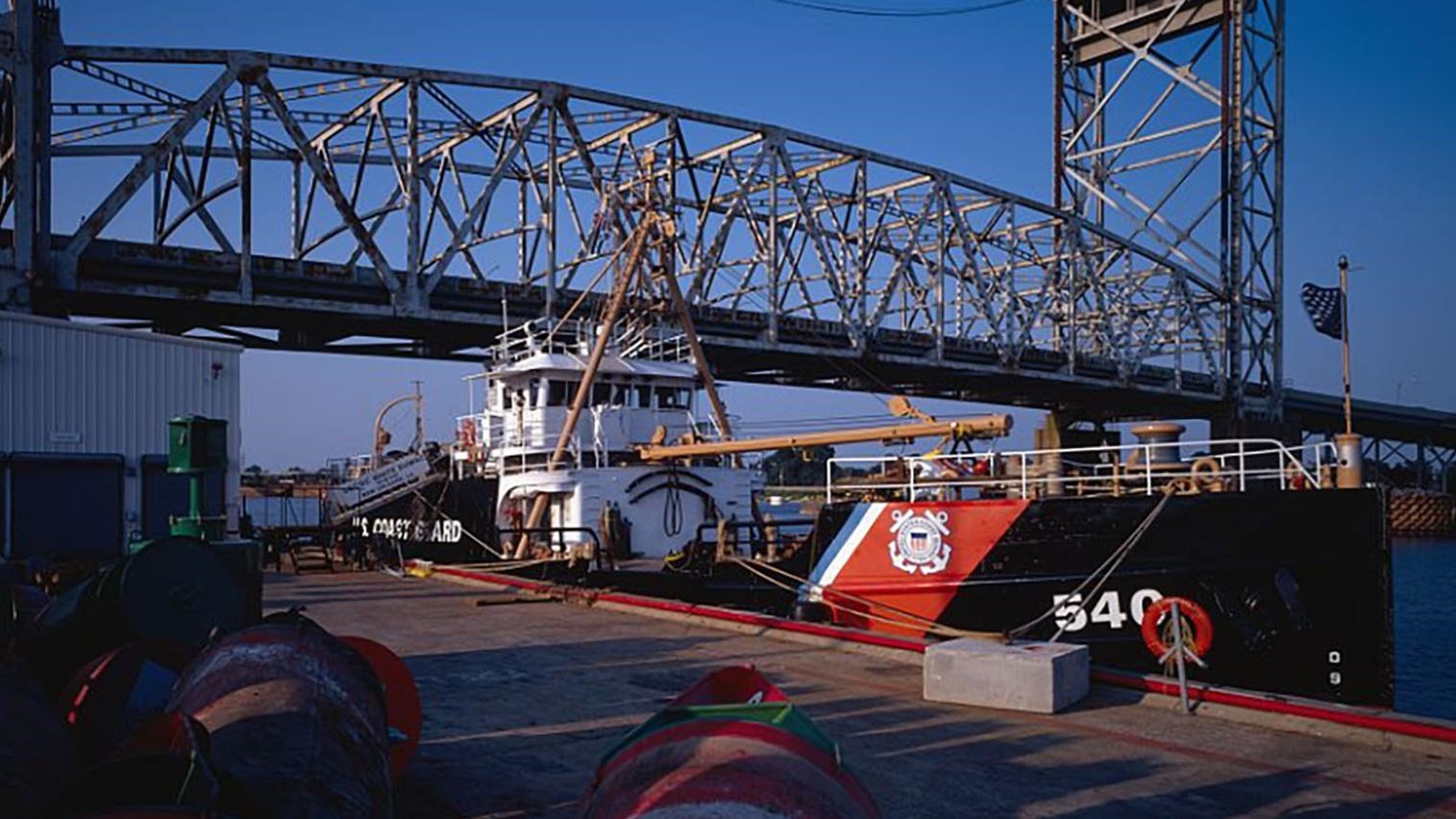 A U.S. Coast Guard Cutter is moored off a concrete pier. A bridge extends above into a blue sky.