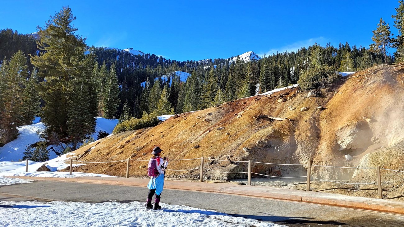 A woman on showshoes takes a photo of a large mudpot in a mountain landscape.