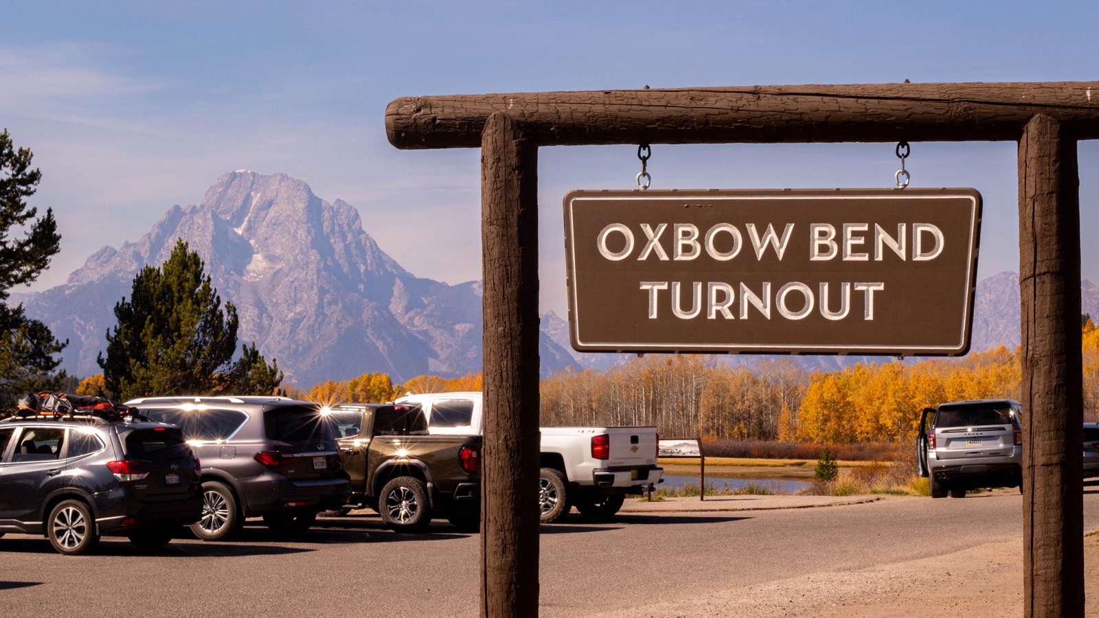 A sign saying Oxbow Bend with cars parked in the foreground and Mount Moran in the background.
