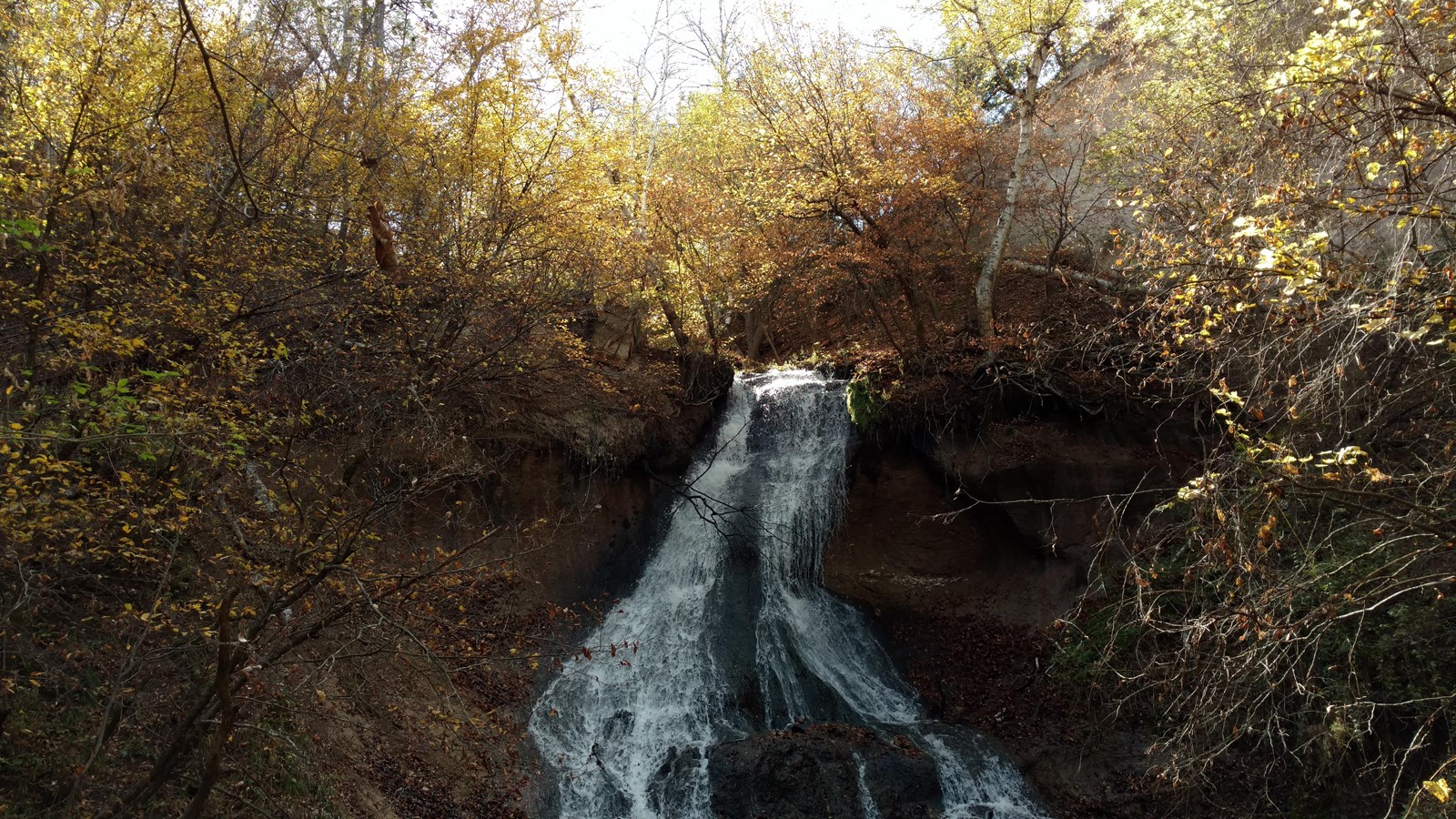 A waterfall flows over a ledge into a canyon in autumn when the leaves are orange and brown.