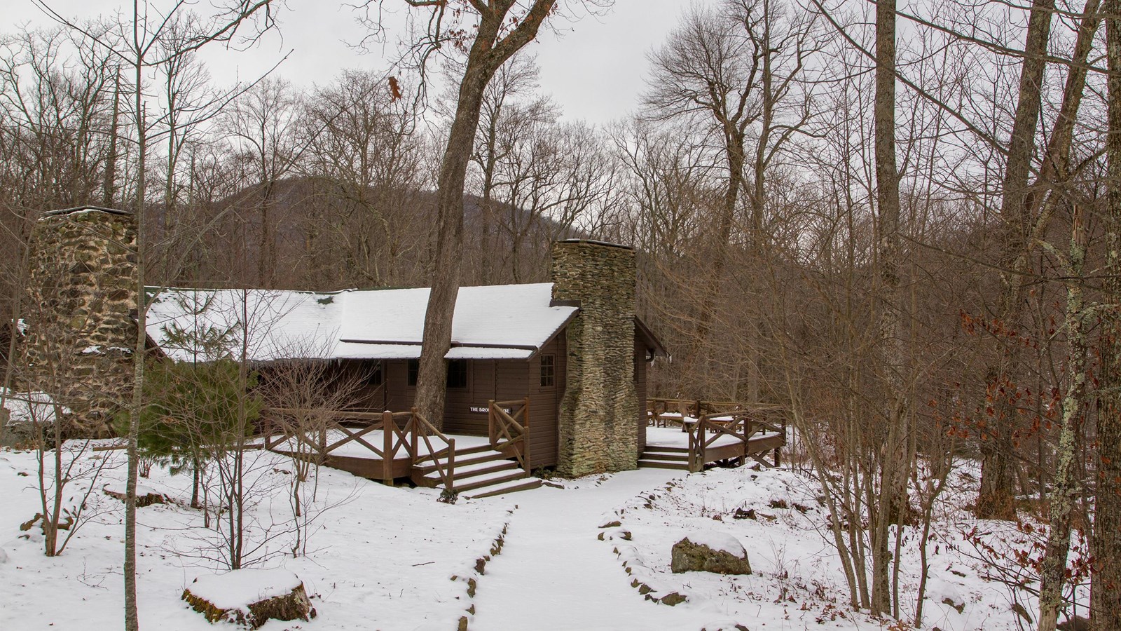 An historic cabin with a large porch sits in the middle of a snow-covered forest.