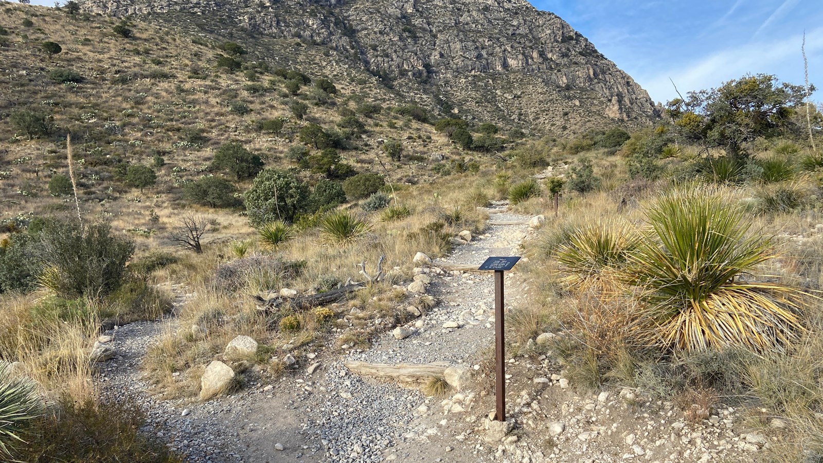 A trail sign marks the junction of three trails in a desert mountain landscape