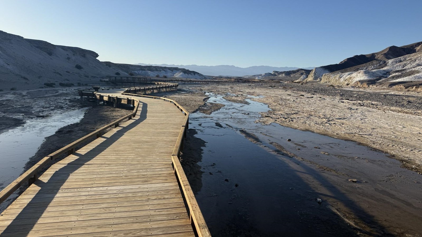 A wooden boardwalk winds back and forth over shallow water in a desert wash with steep banks.