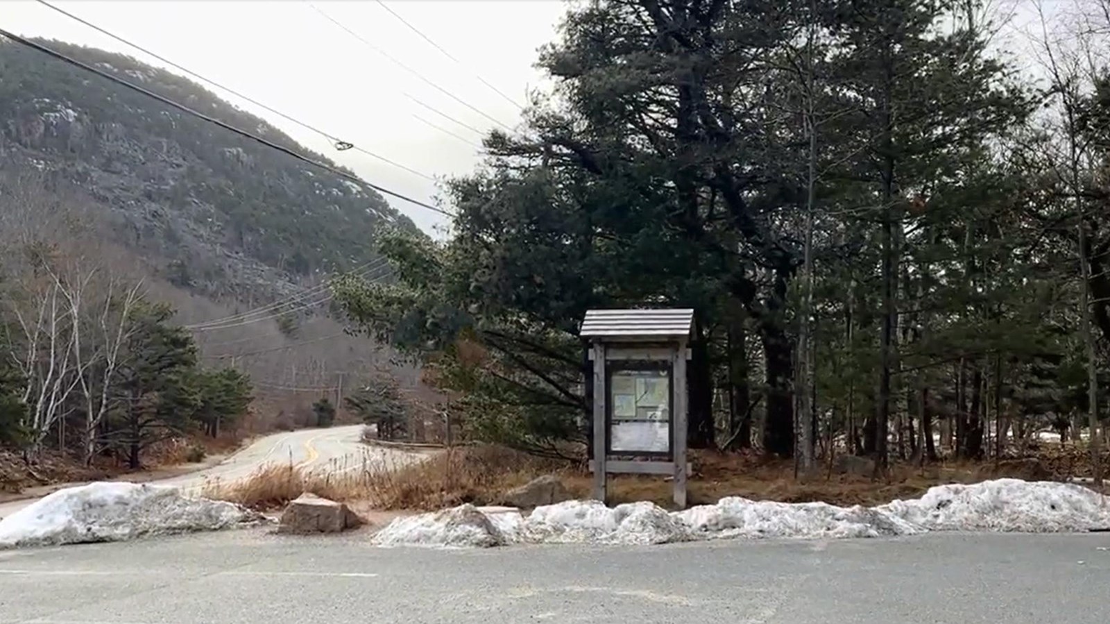 Asphalt parking area along a two-lane highway with a wood trailhead and a steep mountain in distance