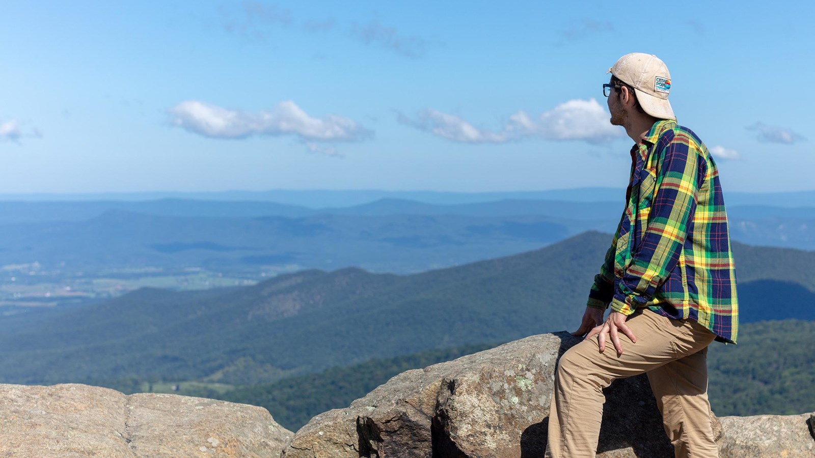 A man in a green shirt and backwards ball cap gazes out over a valley below from a mountain top.