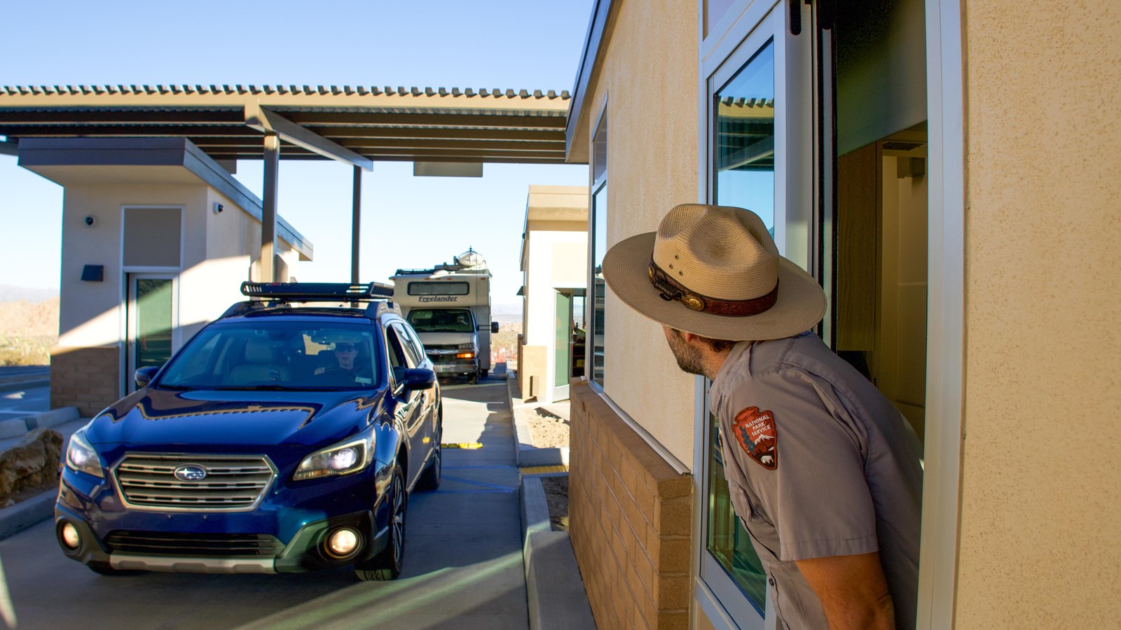 A ranger leans out a fee booth window, while cars pull up to the booth